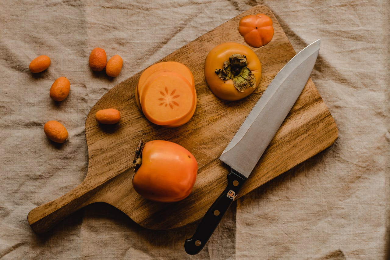 Flat lay of sliced persimmons and kumquats on a wooden board with a knife, showcasing fresh ingredients.