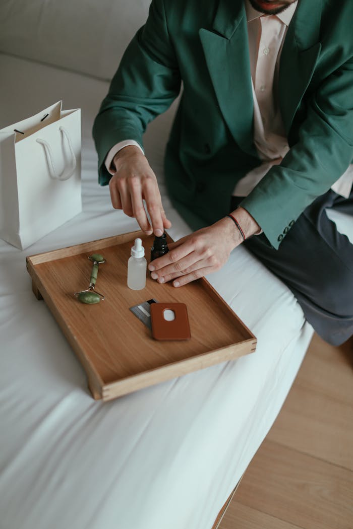 WOODIRE High angle of crop anonymous male in fashionable outfit sitting on comfortable bed and testing various cosmetic products after online shopping