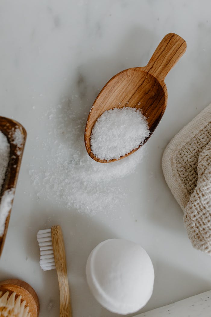 WOODIRE Flat lay of bath salt, wooden scoop, brush, and towel on a marble surface for spa theme.