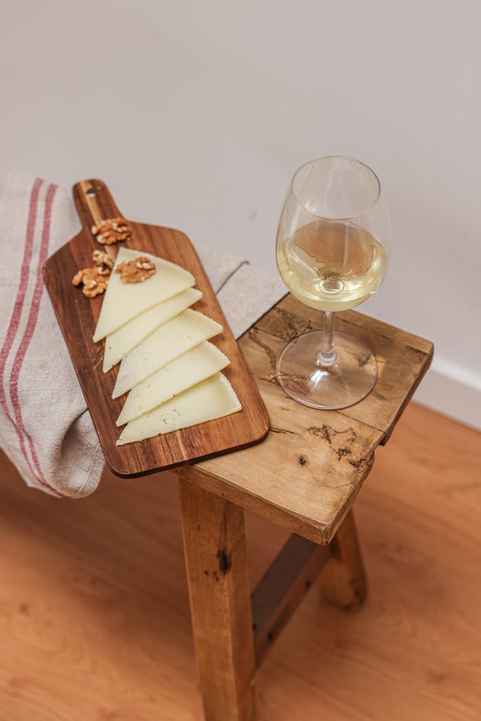 Slices of cheese and walnuts on a wooden board paired with a glass of white wine, creating a cozy indoor setting.